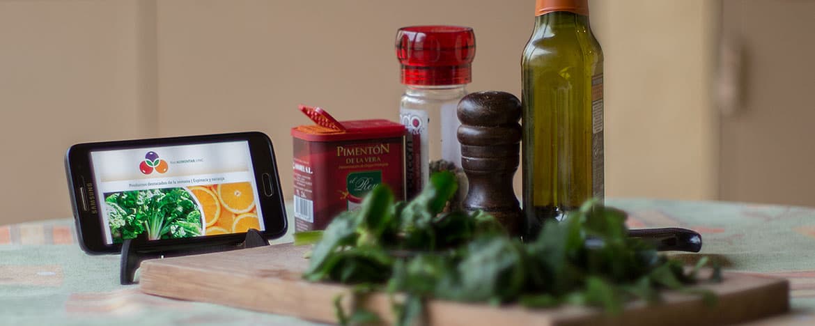 Un celular sobre una mesa junto a una tabla con espinaca, un pimentero, aceite, pimentón y sal