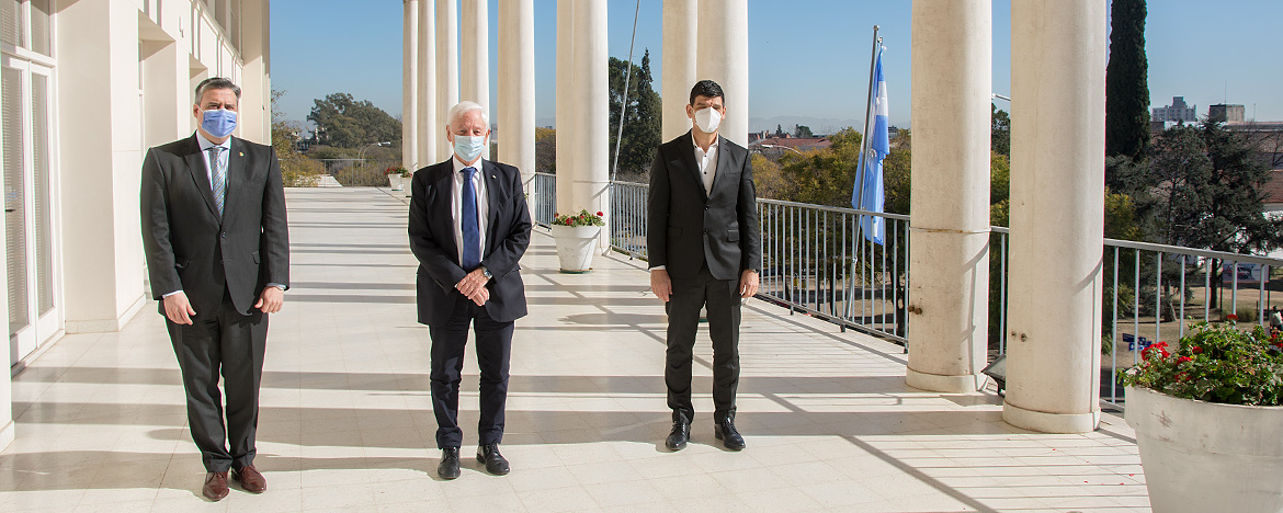 En un día soleado, en un balcón del Pabellón Argentina el rector Hugo Juri, vestido de traje, se encuentra de pie junto al decano de la Facultad de Ciencias Médicas, Rogelio Pizzi, y el secretario de Calidad en Salud de la Nación, Arnaldo Medina, también vestidos de traje